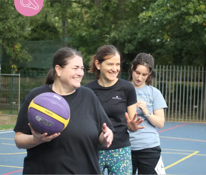 Three women laughing on an outdoor court, one holding a purple basketball